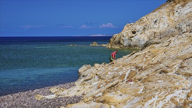 A man climbing on rocks on a coast with clear sky and clear water, Lambi beach, pebble beach,