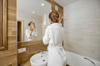 Woman thoroughly washing her face with skin cleanser while standing in a bathrobe in front of a