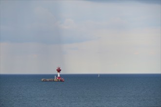 A red lighthouse stands on a calm, blue sea under a slightly cloudy sky, with a small sailing boat