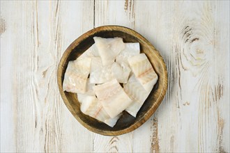 A wooden bowl filled with fresh, cut pieces of white fish fillet sits on a rustic wooden tabletop,