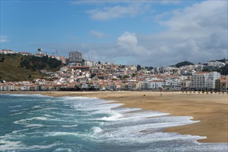 Coastal town with sandy beach, buildings on the hills, and ocean waves under a blue sky, Praia da