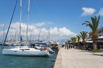 Sailing boats in the harbour next to a promenade under a blue sky with clouds, Mediterranean yacht