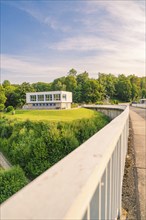 Modern building with large windows on a green hill next to a wide path under a blue sky,