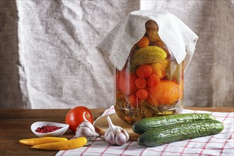 Pickled cucumbers and tomatoes in a glass jar on linen tablecloth and wooden table. Homemade, copy