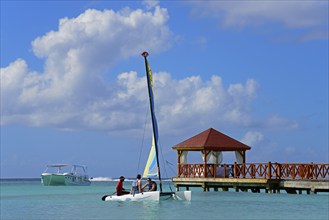 A colourful catamaran sails next to a jetty near a pavilion on the clear sea under a blue sky,