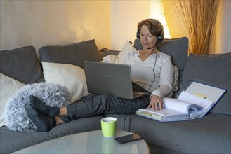 Woman, mid-50s, works from home, with laptop and communicates with colleagues via headset, home