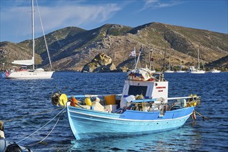 A blue fishing boat in the harbour, surrounded by sailboats and mountains in the background under a