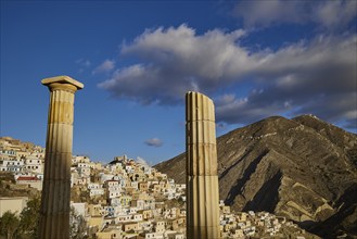 Ancient columns stand in the foreground while a village stretches out in front of mountains,