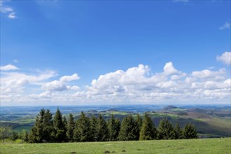 Wide green landscape with mountains and trees under a bright blue sky with white clouds, view from