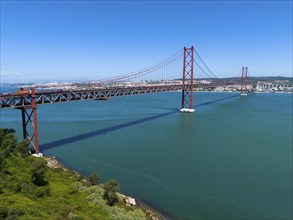 A large red bridge stretches across a wide expanse of water under a clear blue sky, surrounded by