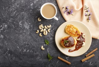 Cheese pancakes with caramel sauce on a beige ceramic plate and a cup of coffee on a black concrete