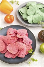 Green and red potato chips with herbs and tomatoes on gray concrete background. Side view, close