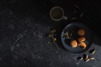 Orange macarons or macaroons cakes with cup of coffee on a black concrete background. Hard light,