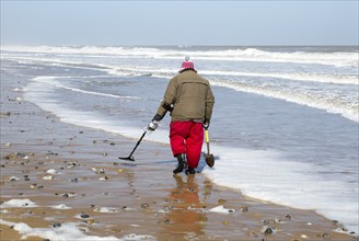 Male detectorist using metal detector walking along sandy beach Hemsby, Norfolk, England, UK