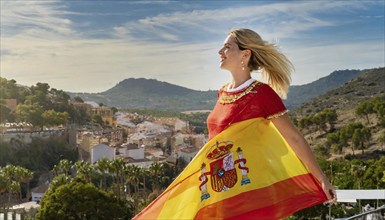 A happy blonde woman holding the flag of Spain, historical town in the background, AI generated, AI