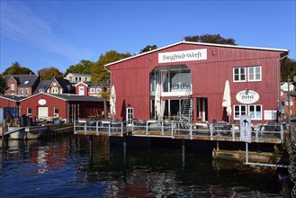 A red wooden building with 'Hotel' lettering on the waterfront under a clear blue sky on a sunny