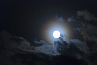 The full moon shines brightly in a cloudy, stormy, black sky and has a halo in rainbow colours