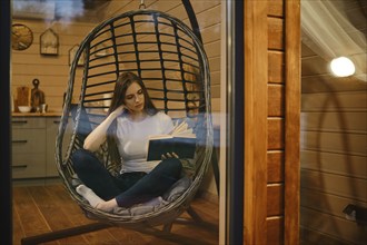 A young woman relaxes in the evening in a wooden house sitting in a hanging chair with a book