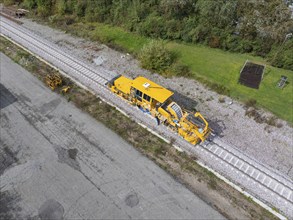 A yellow construction machine working on railway tracks near a road and trees, tamping machine,