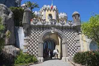 Entrance gate of a castle with decorative stone patterns and plants, visitors are visible, Palácio