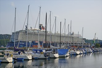 A large cruise ship, Mein Schiff 6, anchored in a quiet harbour with several sailboats in the