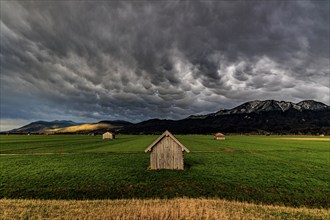 Small wooden hut in a meadow in front of a mountain landscape, storm clouds, thunderstorm, evening