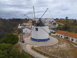 Aerial view of a windmill in the middle of a village, houses with orange roofs and cloudy sky,