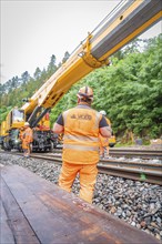 Construction worker in orange safety clothing stands on tracks while a yellow crane works,