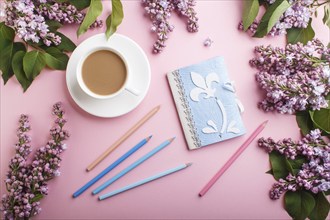 Purple lilac flowers and a cup of coffee with notebook and colored pencils on pastel pink