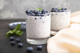 Yogurt with blueberry and chia in glass on black concrete background and orange linen textile. Side