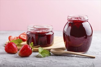 Strawberry jam in a glass jar with berries and leaves on gray concrete background. Homemade, close