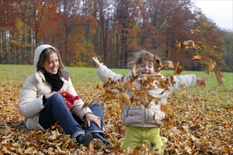 Mother and daughter sitting in a park, daughter playing with autumn leaves, Federal Republic of