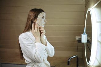 Woman applying sheet mask with mineral elements on her face in front of a mirror