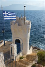 White tower on the coast, with Greek flag in the wind, at blue sea level, Church of Agios Nikolaos