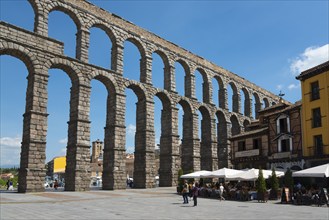 Historic stone aqueduct in a town square with buildings, tables and people, Aqueduct, Segovia,