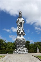 Large stone statue of a majestic figure in front of a blue sky with clouds, surrounded by green