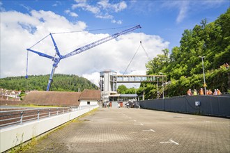 A crane lifts a bridge over a building in sunny weather with blue sky and white clouds, people