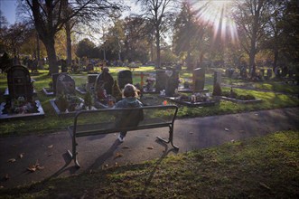 Elderly woman sitting on bench, looking at grave, graves, Tauer, loneliness, autumn colours,