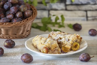 Plum cake on a plate next to a basket with fresh plums in the background, on a white rustic table