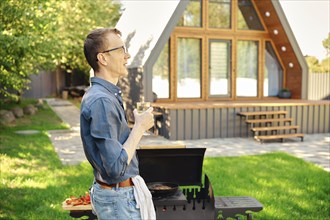 Middle-aged man holds wine glass while standing in profile at the grill in the backyard of a modern