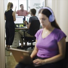 A woman works with headphones on her laptop while colleagues talk loudly. Berlin, 08.08.2024