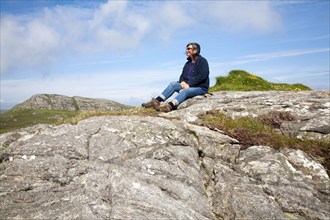 Middle aged female hiker sitting high on rocky outcrop in Barra, Outer Hebrides, Scotland, UK