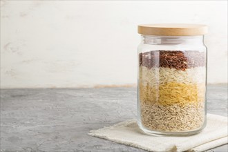 Glass jar with different kinds of rice poured in layers on a gray and white background. side view,