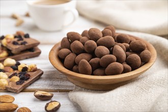 Almond in chocolate dragees in wooden plate and a cup of coffee on white wooden background and