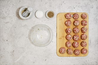 Top view of raw meatballs on wooden cutting board next to empty bowl and spices