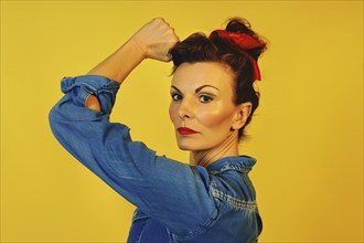 Woman with retro hairstyle and blue blouse raising her hand in fist in front of yellow studio