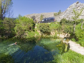 Eine Person schwimmt in einer klaren Quelle in den Bergen neben einem parkenden Van an einem