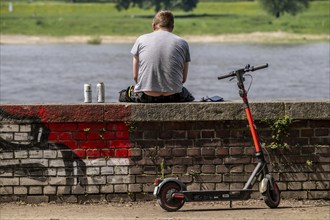 Man sits on a wall, by a river, Rhine, drinks beer from cans, has travelled here on an e-scooter