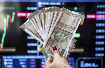 A person holding Indian 500 rupee currency bank note against share market graph on a display