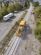 Aerial view of a yellow construction machine on railway tracks with lorries and surrounding green